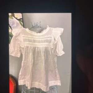 Vit blus med volang och spetsdetaljer - Supersöt vit blus med korta puffärmar, volang vid axlarna och fina spetsdetaljer över bröstet. 