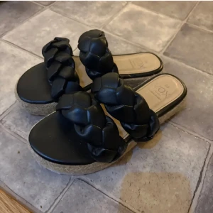 Svarta sandaler med flätade remmar - Snygga svarta sandaler från VOX med två breda, flätade remmar i skinnimitation och plattformsula i kork-look. Perfekta för en trendig sommarstil och riktigt bekväma att glida runt i. Lätta att matcha med både jeans och klänning.