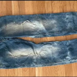 Blå tvättade jeans från Bisko - Säljer ett par blå jeans från Bisko med coolt tvättat och blekt mönster. Jeansen har raka ben och slitna detaljer på låren för en avslappnad streetstyle-look. Perfekta för dig som gillar en unik och trendig vibe.