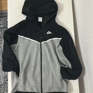 Nike grå och svart tech - Nike vindjacka i grått och svart med huva och hel dragkedja. Jackan har en vit detalj över bröstet, fickor på sidorna och en extra ficka med dragkedja på ärmen. 