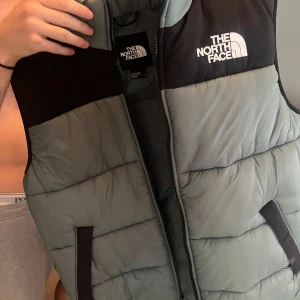 Grå dunväst från The North Face - Säljer en riktigt snygg dunväst från The North Face i grått med svarta axelpartier och vit logga på bröstet. Västen har dragkedja framtill och två fickor med svarta detaljer. Perfekt för lager-på-lager och streetstyle.