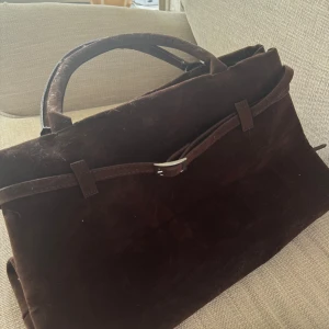 Mörkbrun mocka handväska - Snygg mörkbrun handväska i mocka med stilren design och praktiskt handtag. Väskan har ett spänne framtill och ett rymligt huvudfack med innerficka som stängs med dragkedja. Perfekt för dig som gillar klassiska och tidlösa accessoarer.