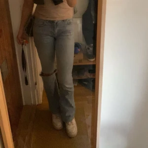 Ljusblå bootcut jeans med hål - Säljer ett par ljusblå jeans med bootcut-modell och ett stort hål på ena knät. Jeansen har klassisk femficksdesign och normal passform. Perfekta för dig som gillar en avslappnad och trendig look.