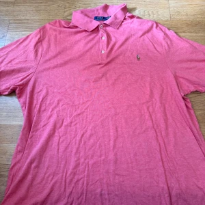 Rosa pikétröja från Ralph Lauren - Snygg rosa pikétröja från Polo Ralph Lauren med klassisk krage och tre knappar framtill. Ikoniska broderade loggan på bröstet. Tillverkad i mjuk bomull som känns skön mot huden. Perfekt för dig som gillar stilren och avslappnad look.