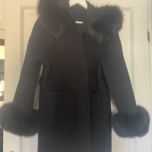 FUR OF STOCKHOLM KAPPA - Snygg svart kappa med stora fickor och fluffiga äktapälsdetaljer vid krage och ärmslut. Rak passform och långärmad modell som håller dig varm under kyliga dagar. (Säljer denna då den inte kommer till användning, nedsatt pris pga att bandet runt tappats)🖤