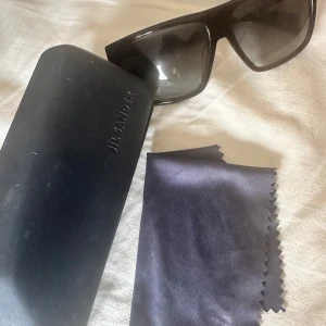 Svarta solglasögon från Jil Sander - Snygga svarta solglasögon från Jil Sander med breda bågar och rektangulär form. Glasögonen har mörka tonade linser och kommer med originalfodral och putsduk. Perfekta för en stilren look och att skydda ögonen mot solen. Finns repa på höger glaset därav priset. Äkta men kommer utan kvitto 
