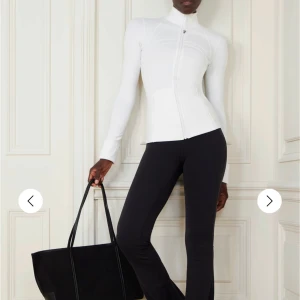 Vit träningsjacka från Lululemon - Slimmad vit träningsjacka från Lululemon med dragkedja framtill, hög krage och långa ärmar med tumhål. Jackan har snygga sömdetaljer och är tillverkad i stretchigt syntetmaterial som andas. Perfekt för gymmet eller powerwalks.