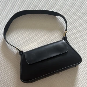 Svart axelväska i skinn med guldspännen - Stilren svart axelväska i slätt skinn med justerbart axelband och snygga guldiga metalldetaljer. Väskan har ett minimalistiskt uttryck med lock och magnetstängning, perfekt för att bära det viktigaste. Enkel att matcha till olika outfits.