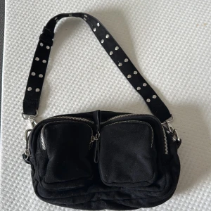 Svart axelväska med nitar och dragkedjor - svart axelväska i mocka med bred axelrem dekorerad med silvriga nitar. Väskan har två stora framfickor med dragkedjor och ett huvudfack, perfekt för att hålla ordning på dina grejer.