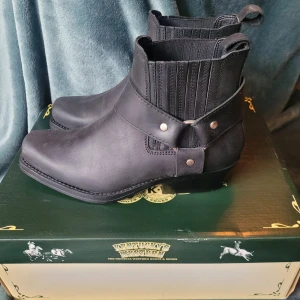 Svarta boots Kentucky's Western - Snygga svarta western boots från Kentucky's Western i skinn med coola detaljer som remmar med nitar och resår vid ankeln. Boots med låg klack och fyrkantig tå, perfekta för dig som gillar western eller bikerstil. Originalpris 1046 kr, slutsåld.  Använda endast en gång.
