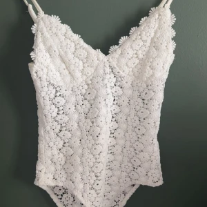 Vit spetsig body i XS från H&M - Superfin vit body från H&M i storlek XS, helt täckt av blommig spets med smala axelband och v-ringning. Toppen har ett romantiskt och somrigt uttryck med detaljerade virkade blommor och fodrad insida för extra komfort.