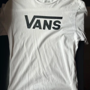 Vit Vans t-shirt med logga - Vit t-shirt från Vans med stor svart logga på bröstet. Klassisk passform och tillverkad i 100% bomull, perfekt för en avslappnad streetstil. Kortärmad modell som är enkel att matcha med jeans eller shorts.