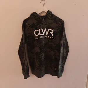 ColourWear hoodie - Snygg hoodie från CLWR i mörkgrönt camouflage-mönster med stor vit logga på bröstet. Tröjan har huva, ribbade muddar och är tillverkad i en mix av ekologisk bomull och polyester. Perfekt för dig som gillar streetwear och vill sticka ut lite extra.