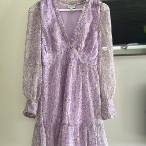 Lila blommig klänning Vero Moda XS - Supersöt lila blommig klänning från Vero Moda Aware. Klänningen har v-ringning, långa transparenta ärmar och volangdetaljer. Den är kort i modellen och har ett romantiskt blommönster i lila och grönt. Perfekt för vår och sommar! Samt för midsommar, någons student osv. Klänningen säljs på grund av att den inte passar mig längre, den är i bra skick, ända är att den är lite uppspred vid V-linjen (Bild 3). Detta är dock inget som syns eller något som påverkar på något sätt. 
