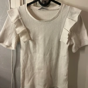 Vit ribbad topp med volang från Zara - Supersöt vit ribbad topp från Zara med korta ärmar och volangdetaljer på axlarna. Den har rund halsringning och är gjord i ett mjukt, stretchigt material! 