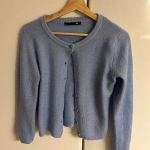 Ljusblå stickad kofta från 157 - Supersöt ljusblå stickad kofta från 157 med rund halsringning och knappar framtill. Perfekt att slänga över en topp eller klänning. Långärmad och mjuk, passar dig som gillar en chill och stilren look. Inte fri frakt! 