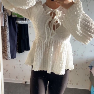 Vit blus med knyt och volang - Supersöt vit blus med volangig nederdel och puffiga långärmar. Toppen har en strukturerad, krinklad yta och snygga knytband framtill vid halsen. Perfekt för dig som gillar romantisk och trendig stil med extra detaljer. Köpt för 500. 