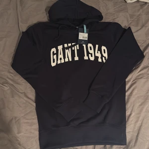 Mörkblå hoodie GANT 1949 XS - Snygg mörkblå hoodie från GANT i storlek XS med vit tryckt text 'GANT 1949' på bröstet. Klassisk huva med snörning och mjuk insida. Perfekt för chill dagar och streetstyle. Materialet är bomullsmix som känns skönt mot huden.