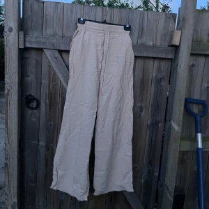 Beiga vida linne byxor med snörning - Säljer ett par beiga linne byxor med breda ben och hög midja. Byxorna har resår i midjan och snörning framtill för extra komfort. Det går att vika ner för att ha lågmidjat. De är tillverkade i ett lätt och luftigt material, perfekta för varmare dagar. Enkel och stilren design som är lätt att matcha. Storlek S men tror skulle passa bättre på storlek XS