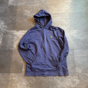 Blå hoodie från Lyle & Scott - Säljer en blå hoodie från Lyle & Scott med klassisk gul logga på bröstet. Tröjan har huva, känguruficka och långa ärmar. Tillverkad i mjuk bomull, perfekt för chill dagar. Enkel och stilren design som funkar året runt.