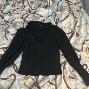 Svart volangtröja från ONLY XS - Supersnygg svart tröja från ONLY med volangdetalj vid halsen och långa ärmar. Tröjan är ribbad vid ärmsluten och har en klassisk, lite elegant look. Perfekt för dig som gillar stilrena plagg med en twist. Tröjan har inga defekter och nästan inte använd. DM vid intresse💕