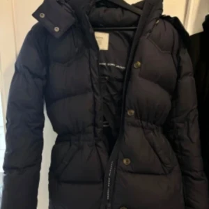 Svart dunjacka från Boomerang S - Snygg svart dunjacka från Boomerang med huva och markerad midja. Jackan har knappar och dragkedja framtill, quiltad design och långa ärmar. Perfekt för kalla dagar när du vill hålla stilen och värmen. Inga repor behöber tvättas och användas 