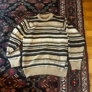 Randig stickad tröja - Säljer en stickad tröja från Co(a)zy i storlek L, passar mer som medium. Tröjan har ett snyggt randigt mönster i beige, svart och grått. Den har rund halsringning, ribbade muddar och är långärmad. Perfekt för dig som gillar en chill och klassisk stil.