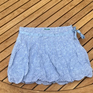 Blommig blå kjol från Benetton - Supersöt ljusblå kortkjol med vitt blommönster från Benetton. Kjolen har ett glansigt satinband i midjan med rosett och dragkedja i sidan. Luftigt material och lätt rynkad nederkant 