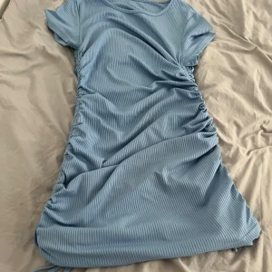 Ljusblå ribbad kortklänning med snörning - Supersöt ljusblå kortklänning med ribbat tyg och snörning längs sidorna som ger en snygg rynkad effekt. Klänningen har korta ärmar och rund halsringning. Perfekt för dig som gillar en enkel men trendig look.