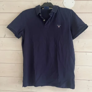 Mörkblå pikétröja från GANT - Snygg mörkblå pikétröja från GANT i klassisk modell med krage och knappar framtill. Tröjan är i bomull och har korta ärmar samt diskret GANT-logga broderad på bröstet. Perfekt för en clean och stilren look.