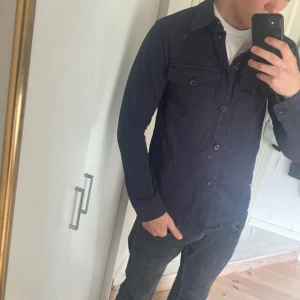 Mörkblå overshirt från Oscar Jacobson - Snygg mörkblå overshirt från Oscar Jacobson med klassisk krage och knäppning framtill. Två bröstfickor med knappar ger en stilren look. Perfekt att bära över en t-shirt för en avslappnad men uppklädd stil.