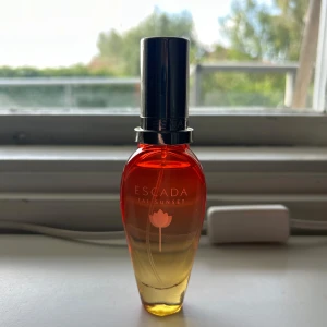 Escada Taj Sunset parfym - Escada Taj Sunset parfym i en snygg glasflaska med färgskiftning från orange till gult och silverfärgad kork. Flaskan har en elegant, smal form och en lotusblomma tryckt på framsidan. Perfekt för dig som gillar fruktiga och exotiska dofter. Säljer pågrund av att jag ej använder.