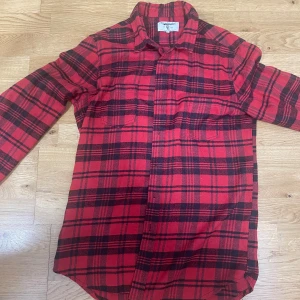 Röd och svart rutig skjorta Cheaploader - Snygg röd och svart rutig skjorta från Cheaploader i storlek L. Skjortan har klassisk krage, långa ärmar och två bröstfickor. Perfekt för dig som gillar en avslappnad och cool stil. Tillverkad i mjuk bomull för extra komfort.