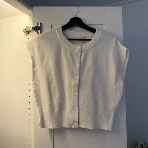 Vit stickad tröja  med knappar - Mysig vit stickad tröja med rund halsringning och knäppning framtill. Tröjan har bred ribbad kant nedtill och vid ärmöppningarna, vilket ger en avslappnad och trendig look. Perfekt att slänga över en skjorta eller t-shirt för extra lager.