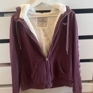 Vinröd zip up hoodie Hollister foder - Mysig vinröd hoodie från Hollister med vit fluffig fleecefoder på insidan. Tröjan har huva med snörning, dragkedja framtill och två fickor. Perfekt för kyliga dagar!