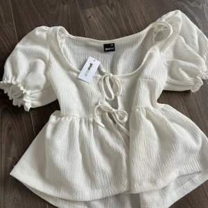 Vit blus med puffärm från Gina Tricot - Supersöt vit blus från Gina Tricot med puffiga korta ärmar och två knytningar framtill. Blusen har en lätt rynkad struktur och en luftig, flowy passform. Perfekt för dig som gillar romantiska detaljer och vill ha en feminin look.
