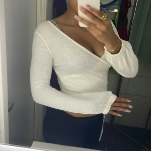 Vit omlottopp med knyt och v-ringning - Säljer en vit omlottopp med v-ringning och långa ärmar. Toppen är croppad och har knyt i sidan för en snygg detalj. Materialet är ribbat och har en lätt struktur som ger en cool känsla. Perfekt för dig som gillar stilrena och trendiga plagg. Har ett litet hål vid armhålan, som lätt man. (Eller jag)  lätt kan sy ihop! Finns ingen storlekslapp men skulle säga att den sitter som s/xs