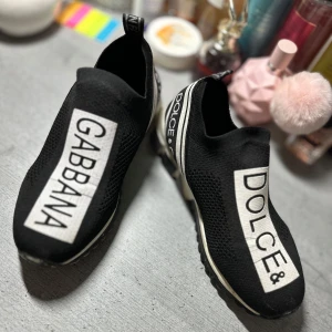 Dolce & Gabbana skor - Svarta slip-on sneakers från Dolce & Gabbana med tydlig logga i vitt framtill och bak. Skorna har en sportig design med stickad ovandel i syntet och elastisk passform. Säljer dem pågrund av att jag och min kille gjort slut de hans gamla som han lämnat kvar, pris e fast försten till kvarn 