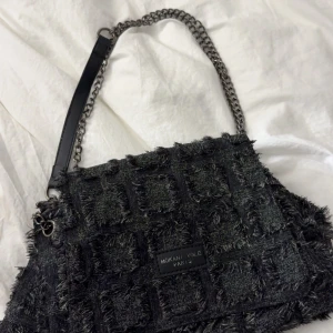 Svart axelväska från Mokani Yole Paris - Säljer en svart axelväska från Mokani Yole Paris med rutigt, fransigt tyg och kedjerem med läderdetalj. Väskan har en cool, edgy look och är perfekt för dig som gillar unika accessoarer. Rymmer allt du behöver till vardags och har ett stort huvudfack.