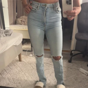 Ljusblå high waist jeans med hål - Säljer ett par ljusblå jeans med hög midja och raka ben. Jeansen har slitna hål vid knäna för en trendig look och klassiska fem fickor. Perfekta för dig som gillar en avslappnad men snygg stil.