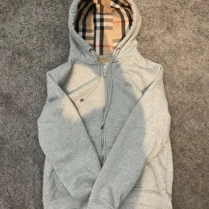 Grå Burberry hoodie med dragkedja - Snygg grå hoodie från Burberry med klassiskt rutigt foder i huvan och broderad logga på bröstet. Tröjan har tyvär problem med dragkedjan där man behöver hålla på med den lite för att kunna dra upp den. Perfekt för dig som gillar stilrena och exklusiva plagg. Storlek M men passar S