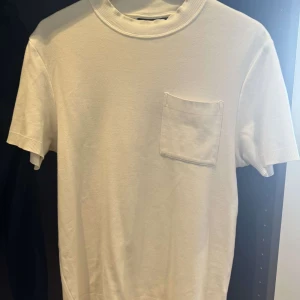 Vit t-shirt j.lindeberg - En stilren vit t-shirt med rund hals och en enkel bröstficka. T-shirten har korta ärmar och ribbade detaljer vid hals och ärmslut. Perfekt för en clean och avslappnad look. Märket är j.lindeberg