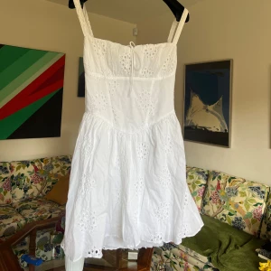 Vit kort klänning med korsett-snörning från GinaTricot - Superfin vit kort klänning i bomull med broderade blommor och snygg snörning i ryggen. Klänningen har breda axelband, söt v-ringning och volangig kjol. Perfekt för sommarens alla tillfällen!