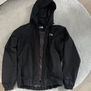 Svart regnjacka från The North Face - Svart regnjacka från The North Face med huva och dragkedja framtill. Jackan har en clean look med logga på bröstet och ryggen, samt elastiska muddar. Tillverkad i slitstark polyester och perfekt för blöta dagar.