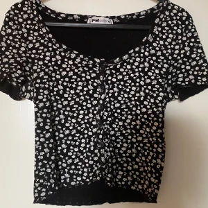 Svart blommig topp från FB Sister XS - Supersöt svart topp från FB Sister i storlek XS med vita småblommor över hela tyget. Modellen är croppad med korta ärmar, rundad halsringning och knappar framtill. Perfekt för dig som gillar en feminin och trendig stil. Frakt tillkommer 