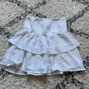 Vit volangkjol med smock - Supersöt vit kortkjol med smockad midja och dubbla volanger. Kjolen har diskret prickigt strukturmönster i tyget och är perfekt för varma dagar. Lätt och luftig känsla, passar till sneakers eller sandaler. Storlek 146/152 men passar xs