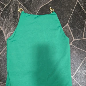 Grön top med guldkedjor xs - Snygg grön klänning med raka linjer och trendiga axelband i guldfärgad kedja. Ärmlös modell som ger en stilren och modern look. Perfekt för dig som vill sticka ut med en cool detalj på axlarna.