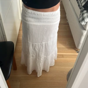 Vit långkjol från Gina Tricot - Vit långkjol från Gina Tricot med smockad midja och lätt, flowigt tyg. Kjolen har en bohemisk känsla och är perfekt för varma dagar. Den är tillverkad i mjuk bomull och har en luftig passform som ger en avslappnad vibe. Använd få gånger, har dock en liten fläck längst ner baktill som knappt syns (bild 3). Storlek 146/152. Mått: 80 cm lång💗