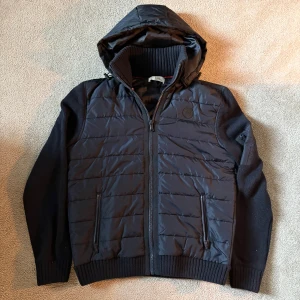 Mörkblå cardigan från Pier One - Mörkblå cardigan från Pier One med luva och ribbstickad krage. Jackan har quiltad framsida och baksida i syntetmaterial, samt stickade ärmar och muddar. Dragkedja framtill och två fickor med dragkedja. 