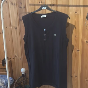 Mörkblå ärmlös topp från Lacoste - Snygg mörkblå ärmlös topp från Lacoste med rund halsringning och tre knappar framtill. Klassisk krokodil-logga broderad på bröstet. Tillverkad i mjuk bomull, perfekt för en clean och sportig look.